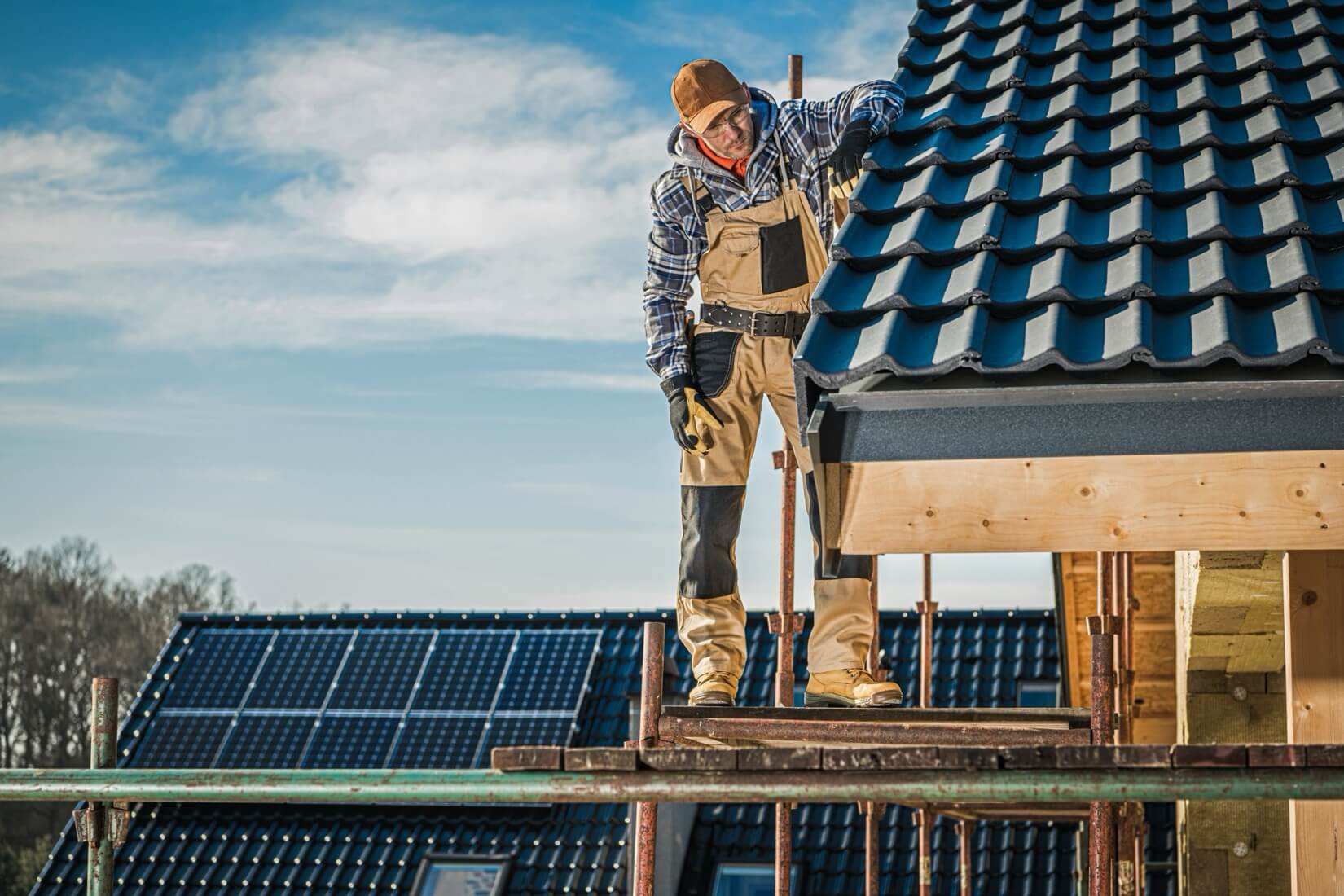 En takläggare står på en byggställning och arbetar med takläggning på ett nytt hus, med solpaneler synliga på ett annat tak.