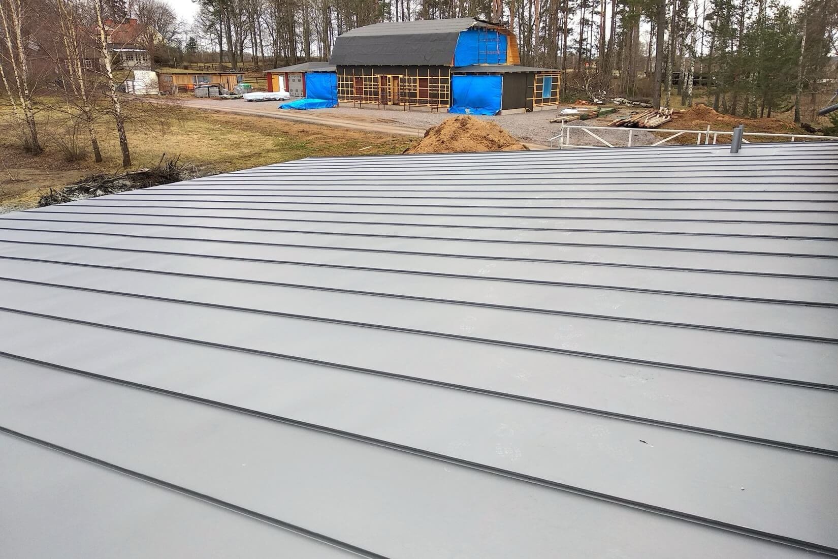 Ett nylagt, grått plåttak med bandtäckning på ett hus, med en annan byggnad under konstruktion i bakgrunden, som visar ett projekt inom takläggning.